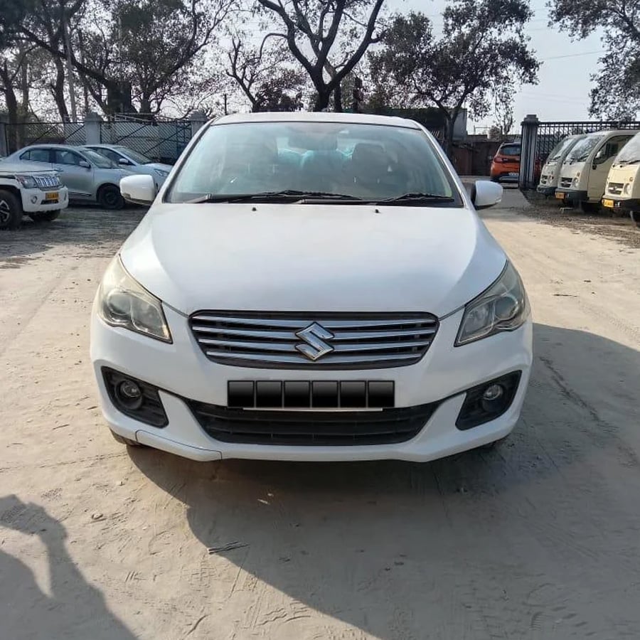 A white Suzuki sedan is parked on a dirt lot, facing forward, with trees and several other vehicles visible in the background on a sunny day.