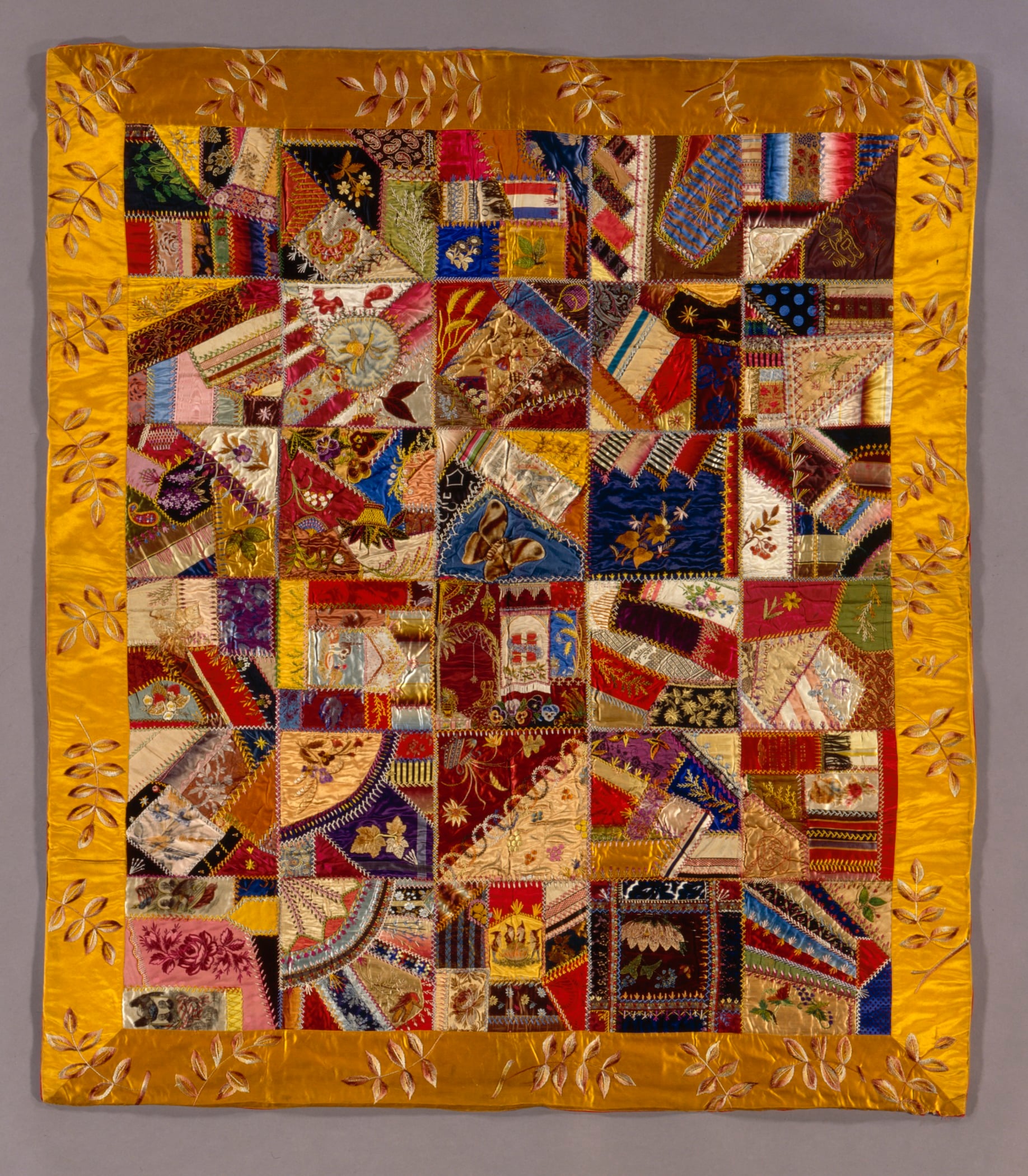 A colorful patchwork quilt with intricate patterns and embroidery, bordered by a gold fabric featuring leaf designs. The quilt consists of irregularly shaped fabric pieces in a variety of colors and textures.