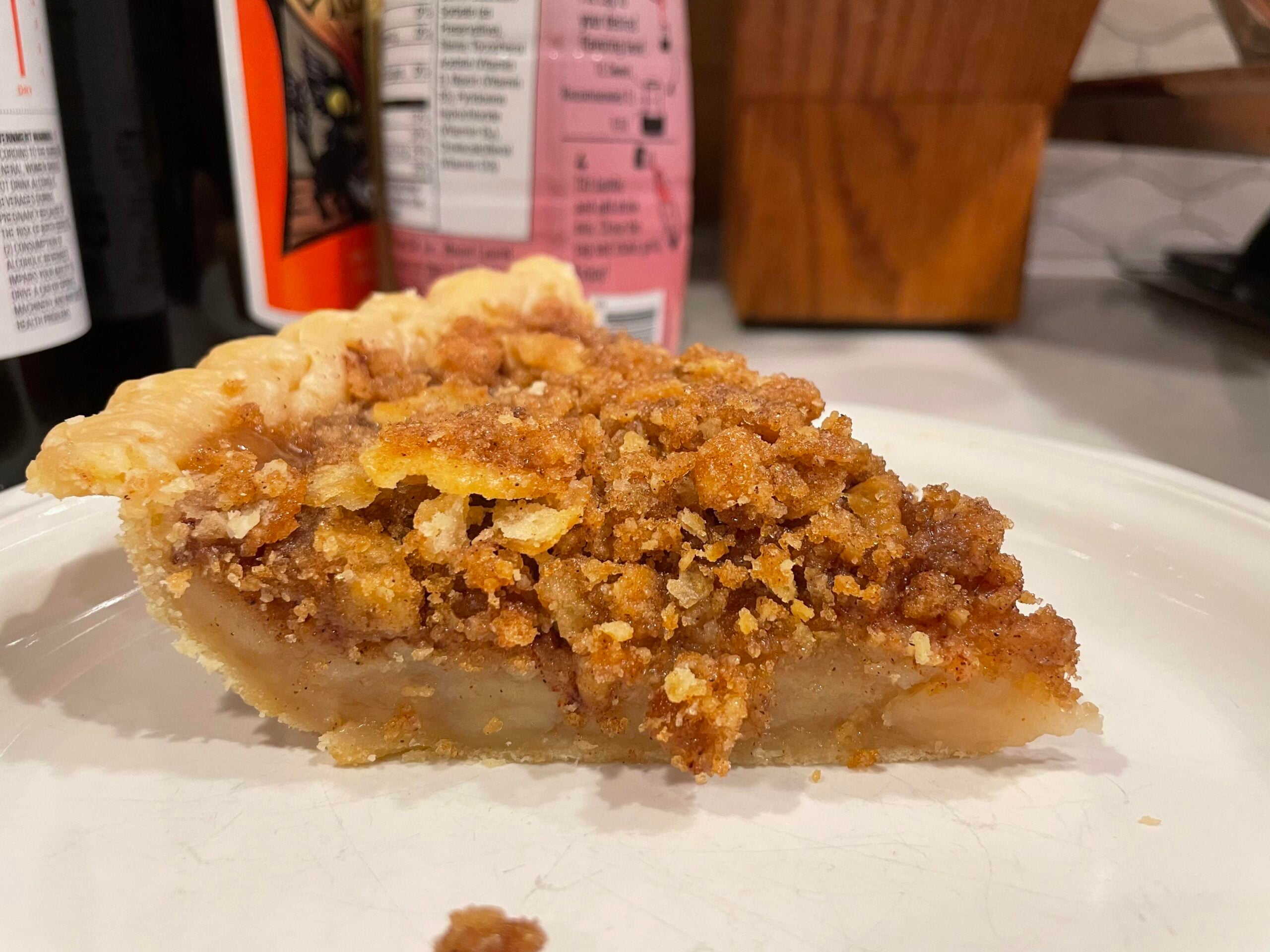 A slice of crumb-topped pie with a golden brown crust and cinnamon filling sits on a white plate, with bottles and wooden objects blurred in the background.