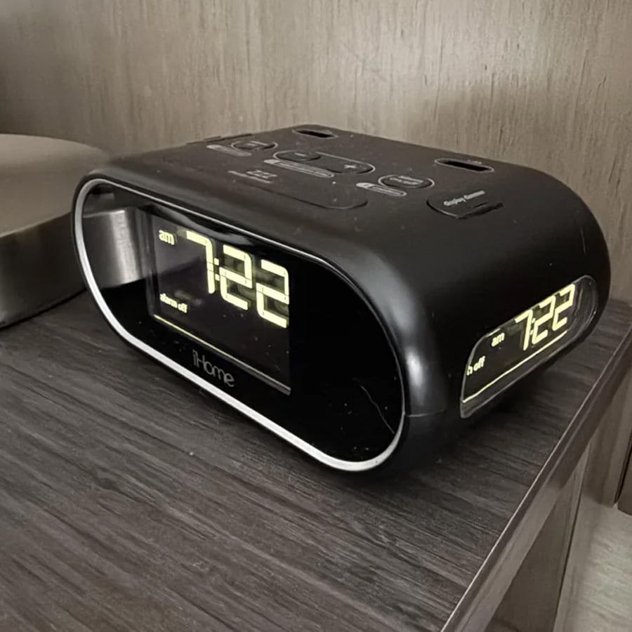A black iHome digital alarm clock displays the time 7:22 on a wooden nightstand, with a wall and part of another object in the background.