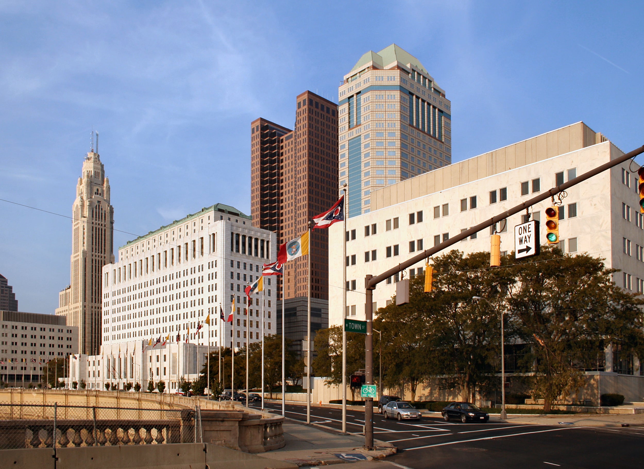 Downtown cityscape with tall buildings, multiple flags, and a traffic light at an intersection on a sunny day. Trees line the sidewalk near the white and beige office buildings.