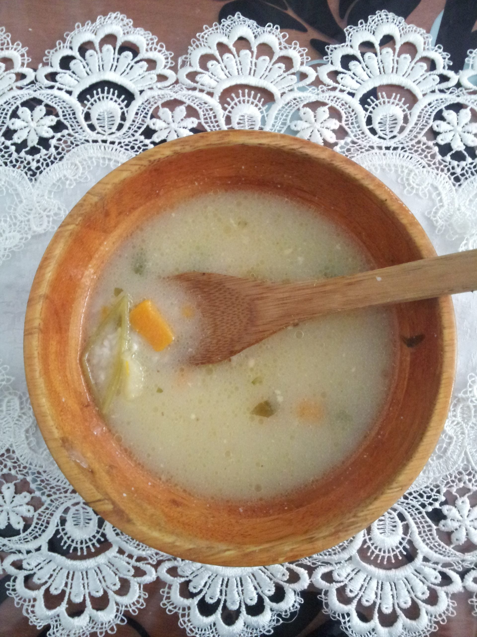 A wooden bowl of creamy soup with visible vegetables and a wooden spoon, placed on a white lace tablecloth.