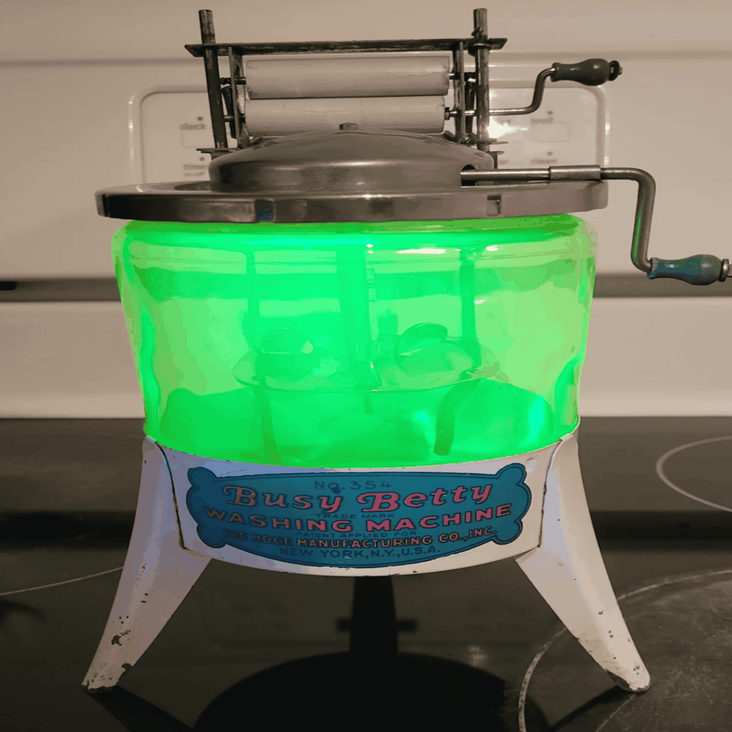 A vintage Busy Betty toy washing machine with a glowing green light inside, metal hand crank, and blue and white label, is displayed on a stovetop.