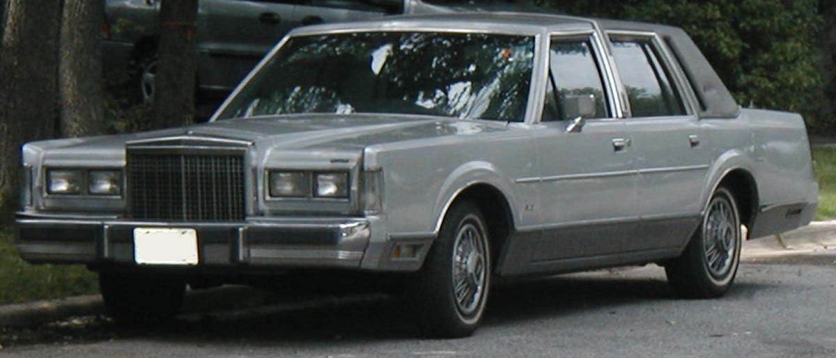 A silver, four-door classic sedan parked on a street, surrounded by trees and greenery, with another car in the background.