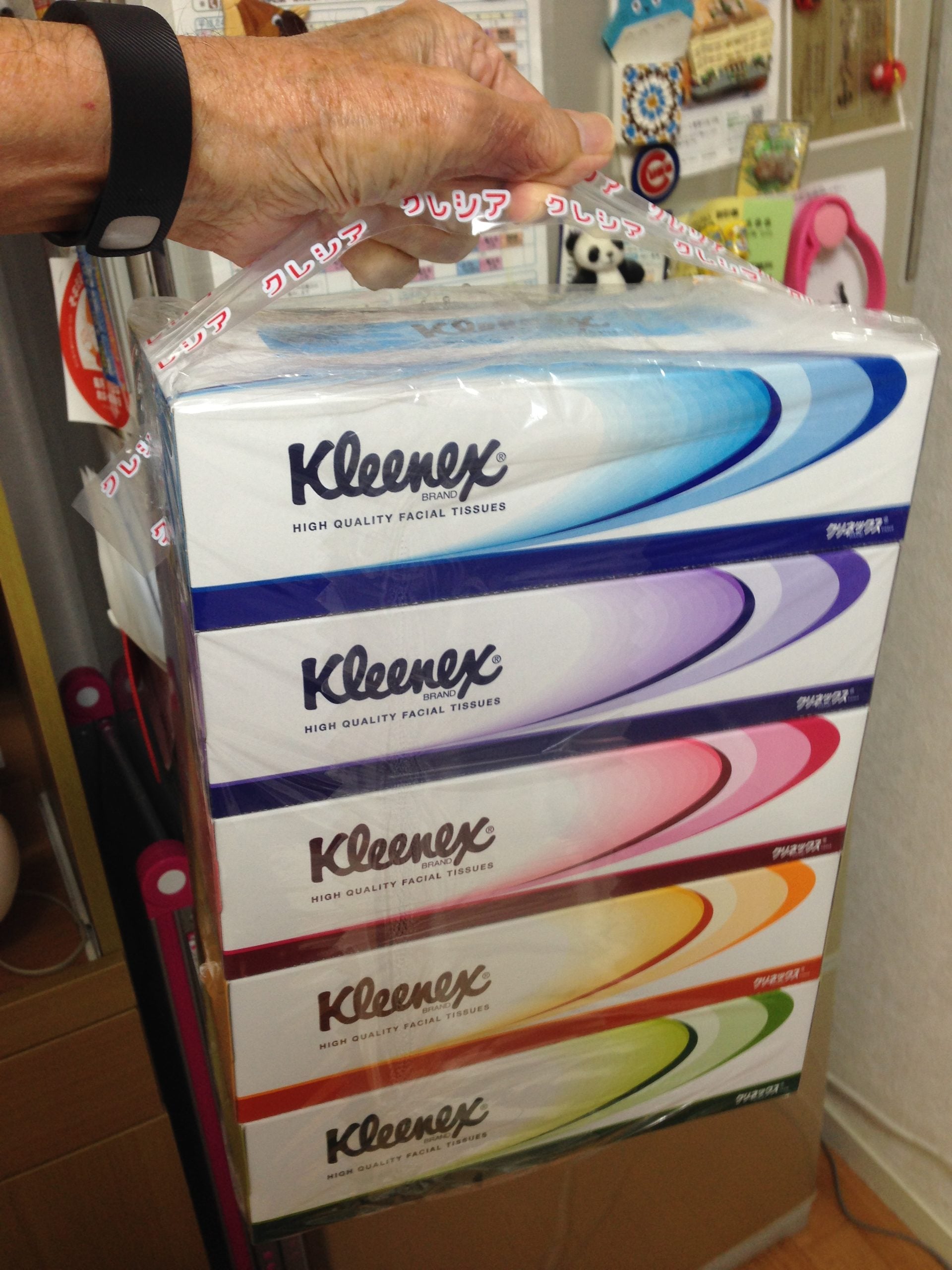A hand holds a pack of five stacked Kleenex tissue boxes, each with a different color swoosh design, in a home setting.