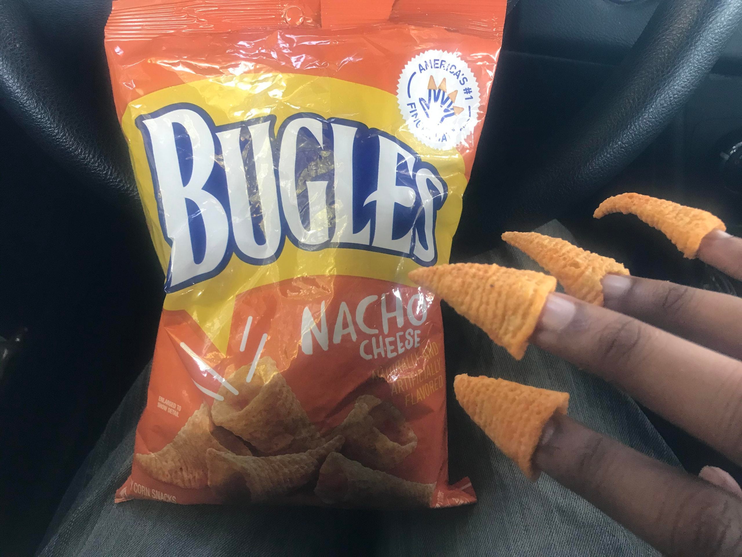 A hand with Bugles snacks on each fingertip is shown in front of an orange bag of Bugles Nacho Cheese flavored snacks.