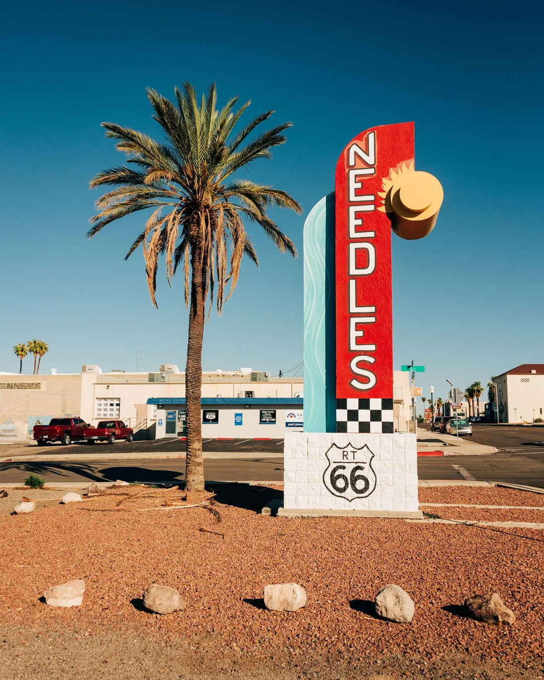 A large retro sign reading “NEEDLES” stands beside a palm tree on Route 66 in Needles, California, under a clear blue sky. Buildings and parked cars are visible in the background.