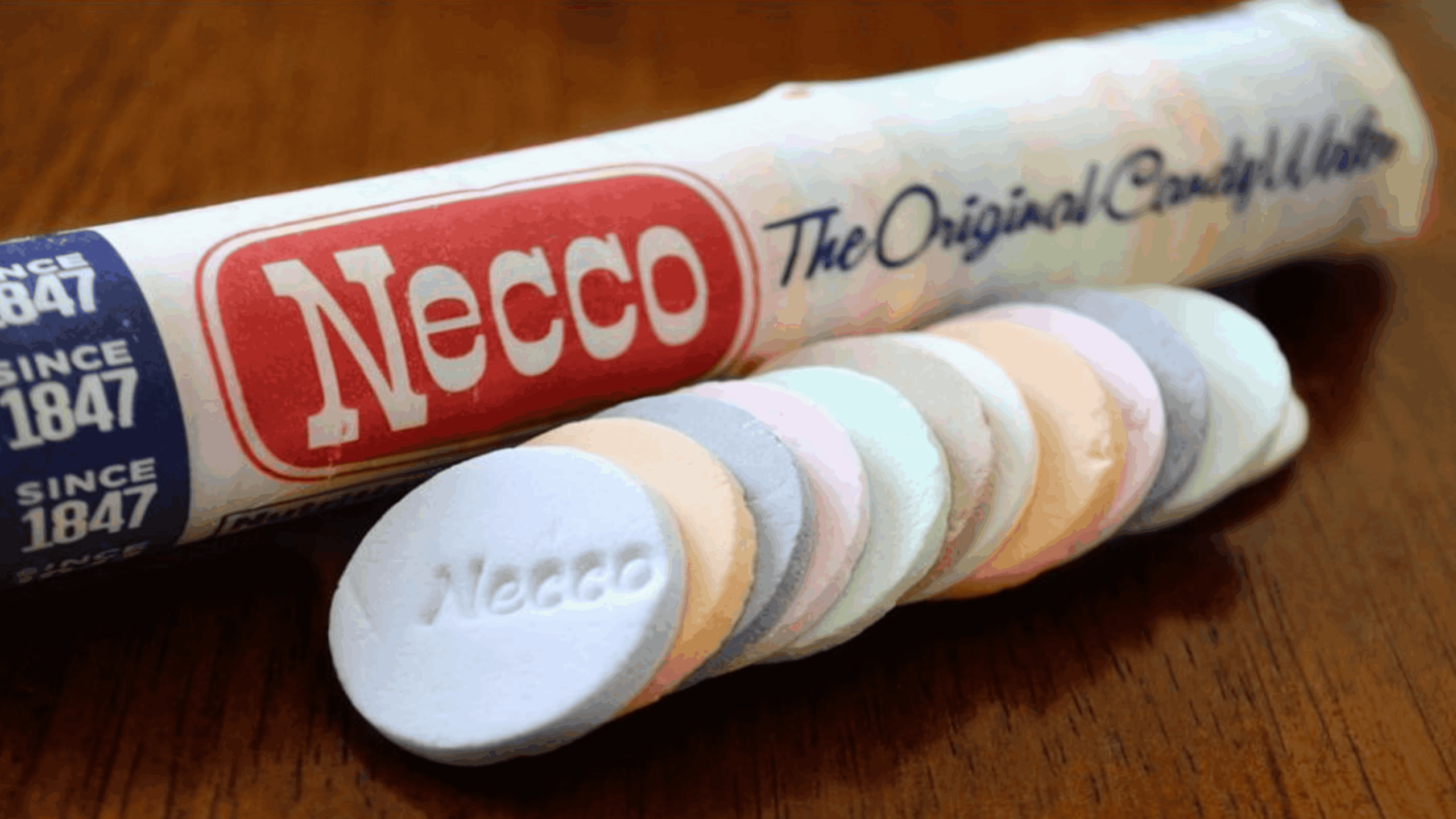 A roll of Necco Wafers with several pastel-colored candy wafers spread out in front, resting on a wooden surface.