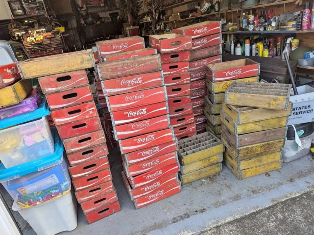 Stacks of vintage red Coca-Cola crates and weathered yellow wooden crates are arranged in a cluttered garage. Shelves with cans, bottles, and storage bins are visible in the background.