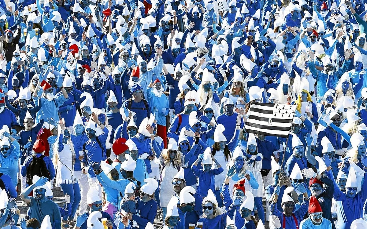 A large crowd dressed as Smurfs, with blue faces, white hats, and blue clothing, gathers outdoors. One person is holding a black and white striped flag. The group appears festive and cheerful.