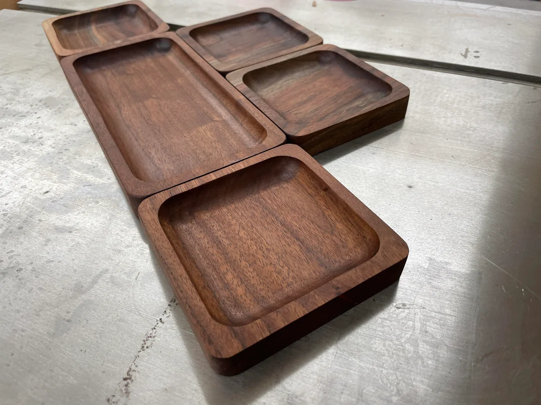Five rectangular wooden trays with rounded edges are arranged on a metallic surface. The trays vary slightly in size and are made of dark wood, possibly walnut.