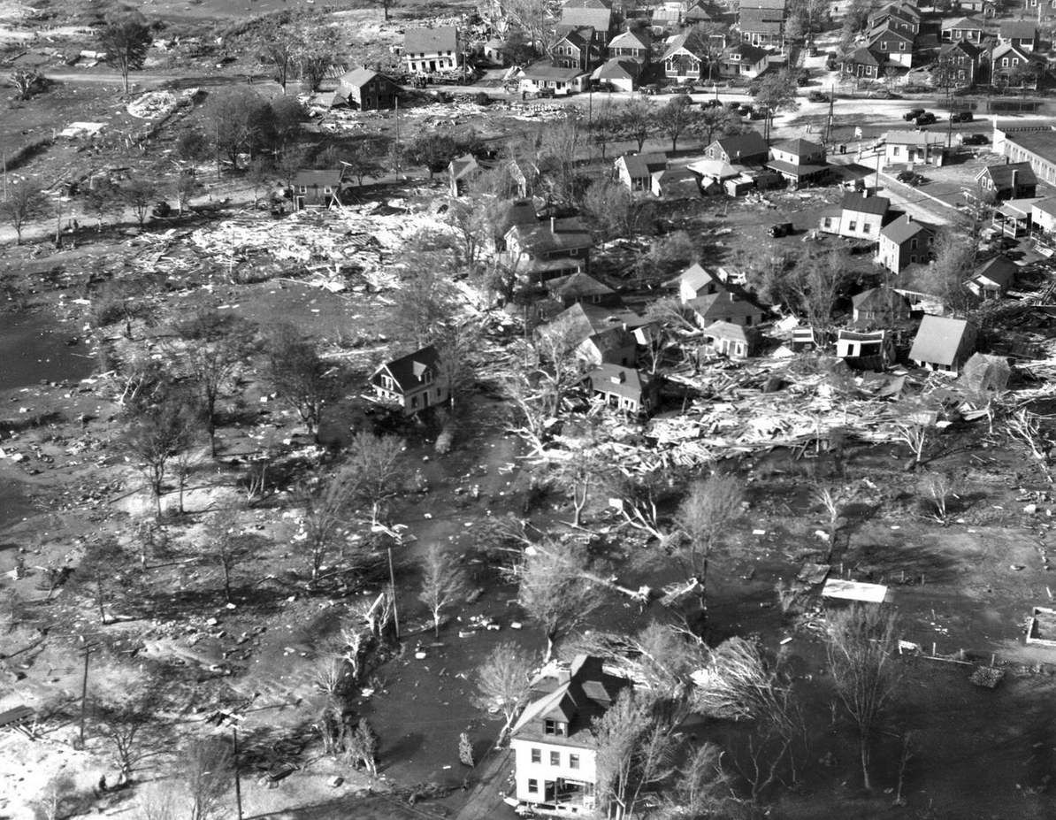 Aerial view of a residential area devastated by flooding, with damaged homes, debris scattered around, and water covering parts of the land. Some houses remain standing while others are heavily damaged or destroyed.