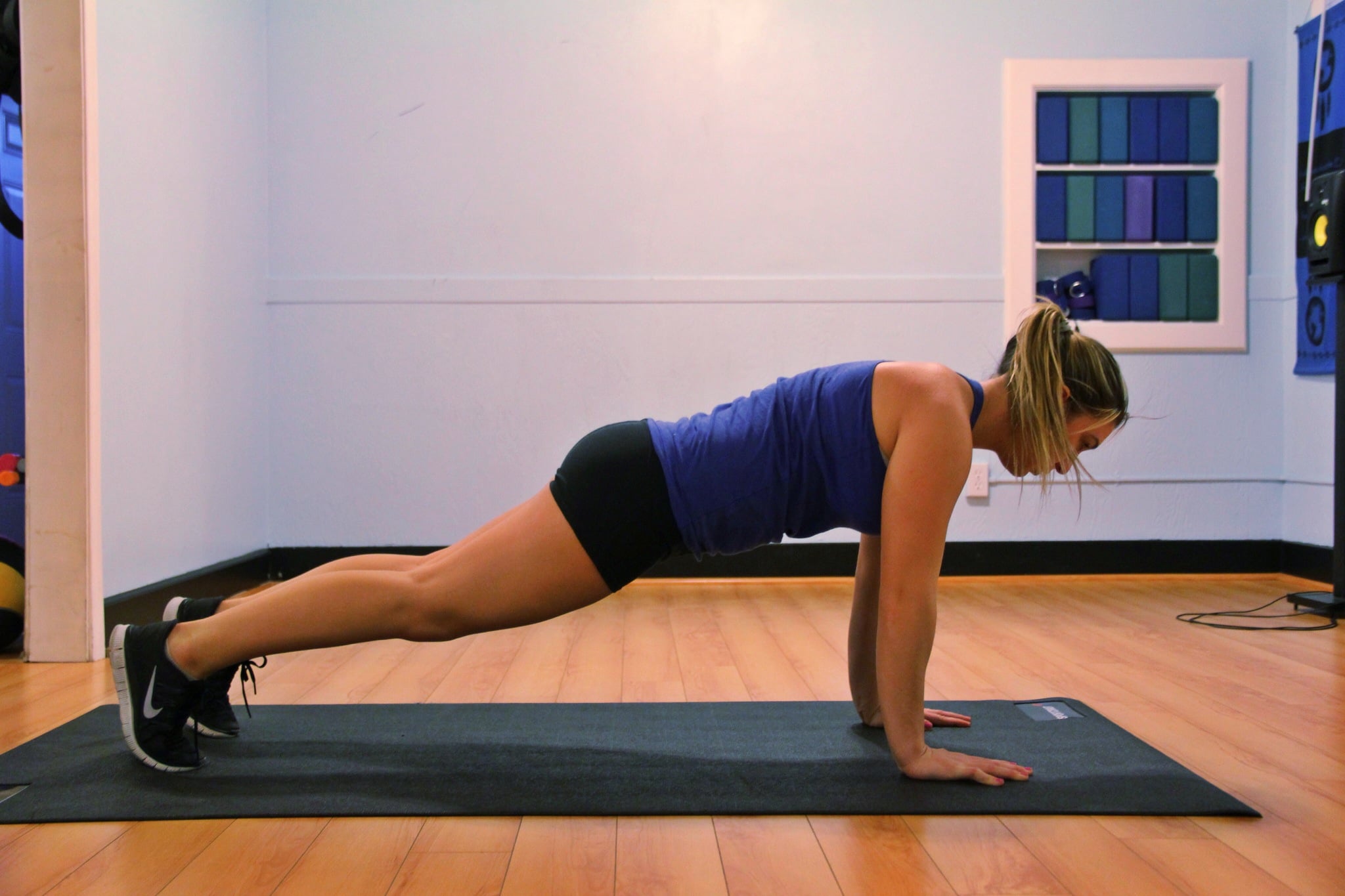 A woman in athletic wear is doing a plank exercise on a yoga mat in a brightly lit room with wooden floors and light blue walls.