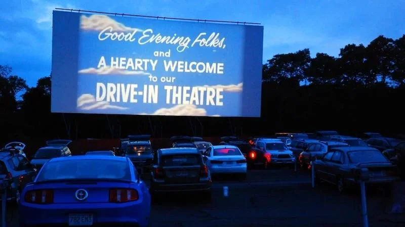 Cars parked at a drive-in theater at dusk, facing a large screen that reads, “Good Evening Folks, and A HEARTY WELCOME to our DRIVE-IN THEATRE,” with trees silhouetted in the background.
