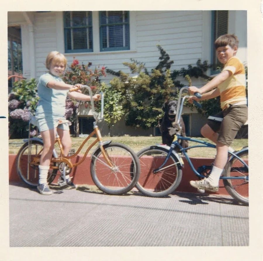 Two smiling children on bicycles pose by a red curb in front of a house, with a black dog sitting between them. Flowers and bushes are visible in the background.