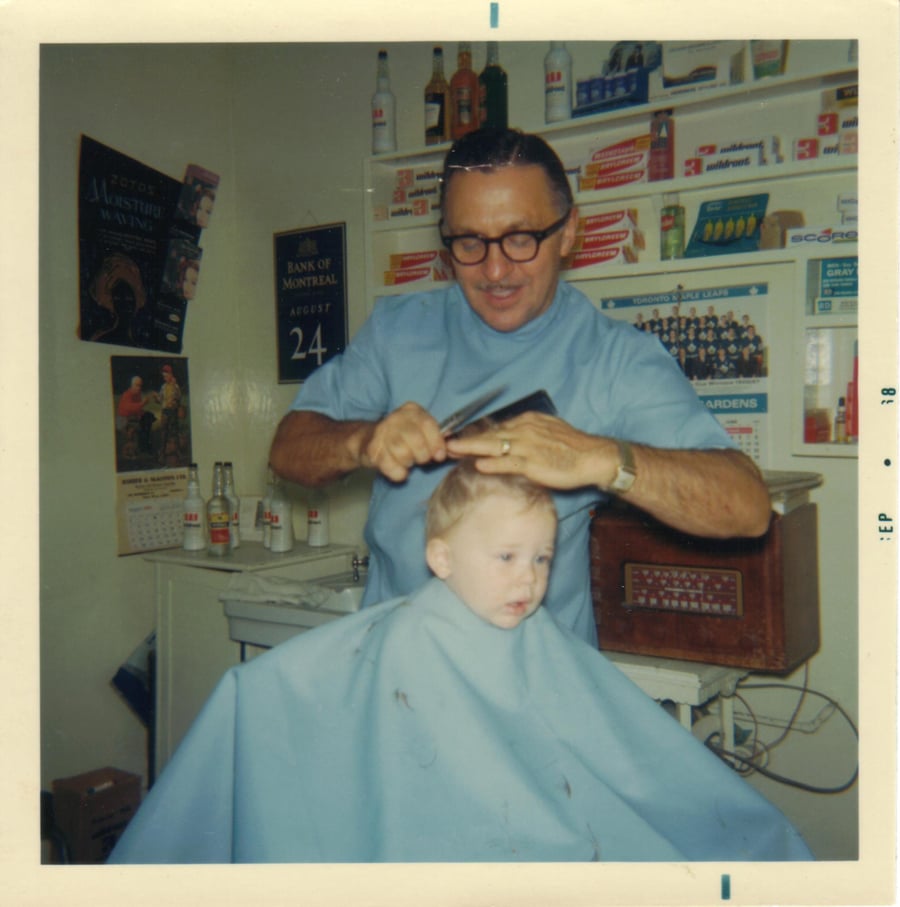 A barber in glasses cuts a young boy’s hair in a vintage barbershop, with shelves of products and soda bottles in the background. The boy is covered in a blue cape.