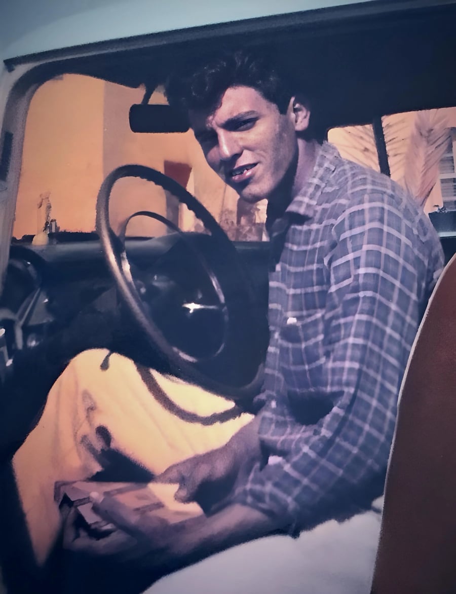 A man in a plaid shirt sits in the driver’s seat of a car, smiling at the camera. He holds something in his hands and appears relaxed. The photo has a vintage feel.
