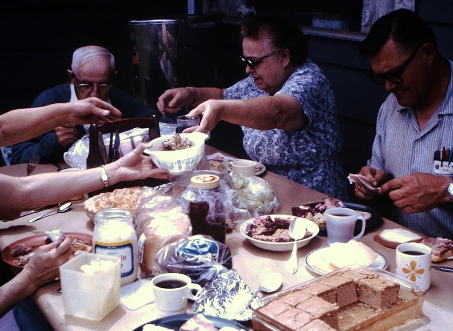 Four adults share a meal outdoors at a table filled with food, desserts, coffee, and jars. One person serves a dish while others eat and reach for items. The scene feels casual and communal.