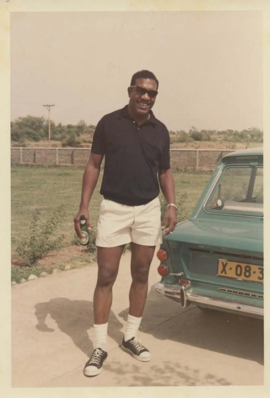 A man wearing sunglasses, a dark polo shirt, white shorts, and black sneakers stands smiling next to a vintage green car, holding a bottle outside on a sunny day.