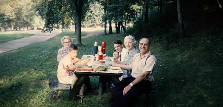 Six people sit at a picnic table in a grassy, wooded park, sharing a meal. Various food containers and drinks are on the table, and everyone is smiling at the camera. Sunlight filters through the trees.