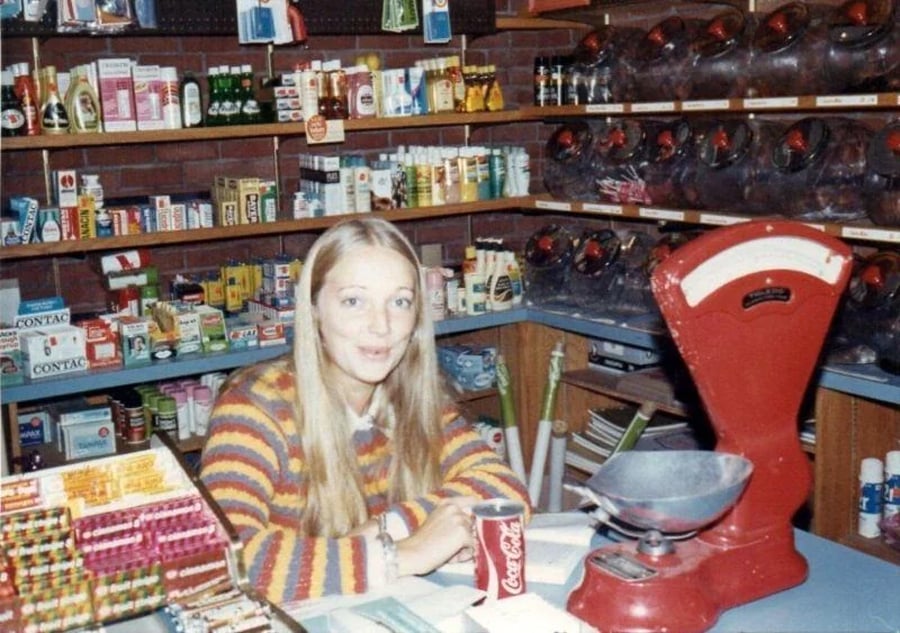 A woman with long blonde hair and a striped sweater sits at a counter in a vintage store, with a red scale, a Coca-Cola can, and various products on the shelves behind her.