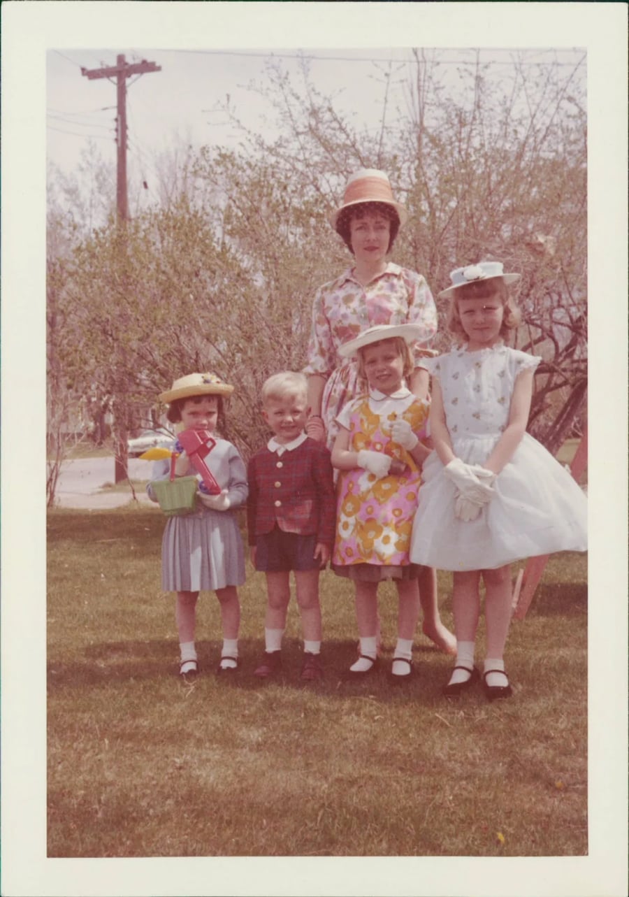 Four children and a woman stand outside on grass, all dressed in colorful spring or Easter outfits with hats. The children hold baskets and wear gloves. Trees and a telephone pole are in the background.
