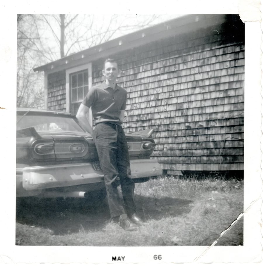 A man stands next to a vintage car in front of a wooden shingle building. The photo is in black and white and dated May 1966. The man is smiling and dressed in a short-sleeve shirt and pants.
