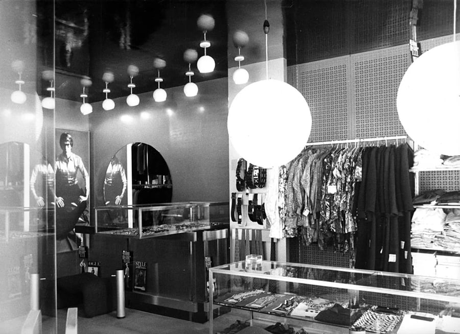 Black-and-white photo of a clothing store with hanging lights, folded clothes on glass counters, a wall rack displaying shirts, and a mirror reflecting part of the store. A large poster of a man is on the left wall.