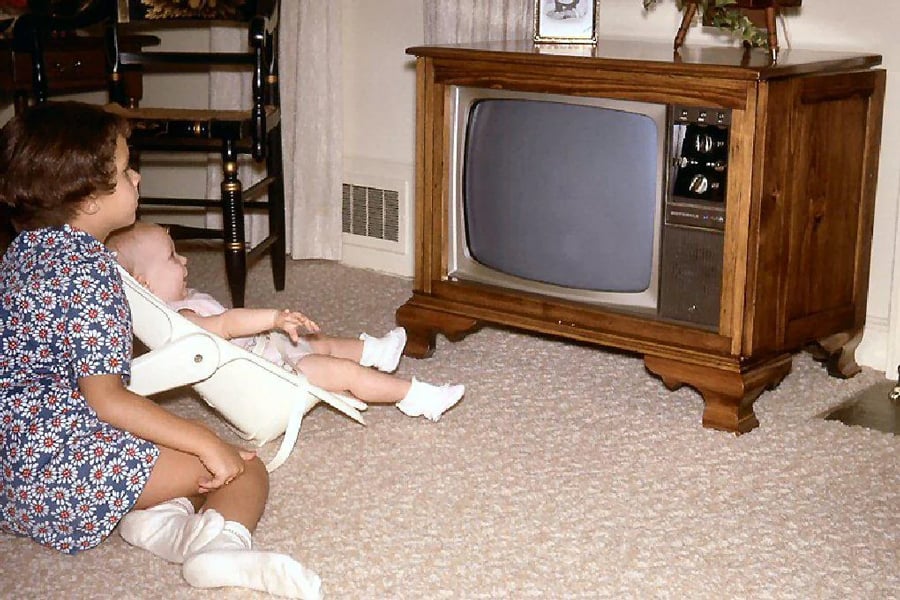 Two children sit on a carpeted floor watching an old-fashioned wooden television; one is in a baby seat and the other sits beside them, both facing the TV in a cozy living room.