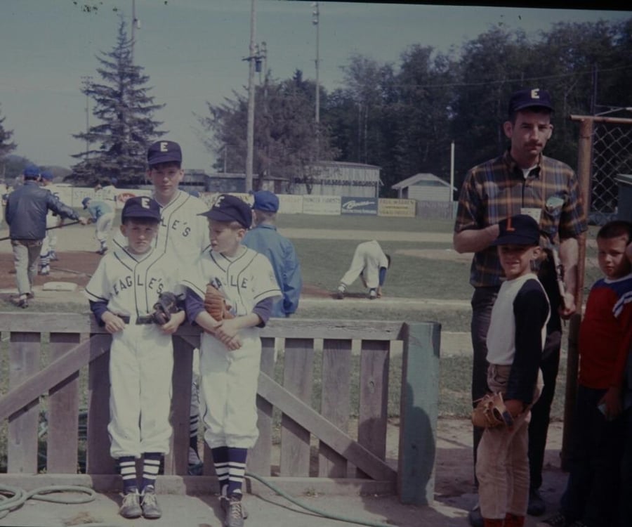 Three boys in Eagles baseball uniforms stand by a wooden gate at a baseball field. An adult and two other kids stand nearby. Players and coaches are visible on the field and in the background.