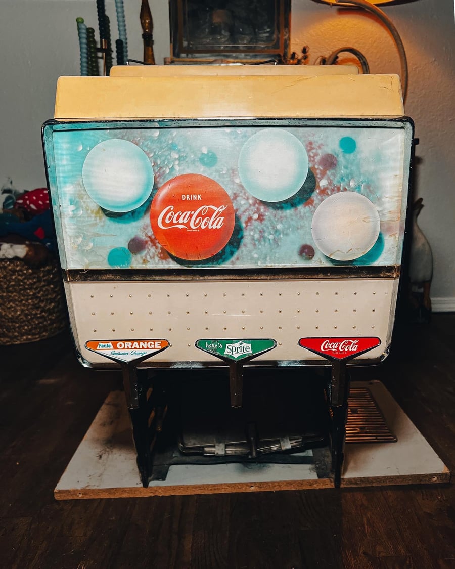 Vintage soda fountain machine with three levers for Fanta Orange, Sprite, and Coca-Cola, featuring a retro Coca-Cola design and worn paint, sitting on a wooden floor indoors.