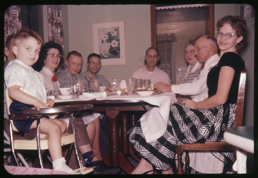 A group of adults and one child sit around a dining table, smiling at the camera. The table is set with dishes and glasses. The setting appears to be a cozy, mid-20th-century home.
