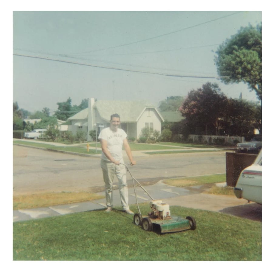 A person is mowing a green lawn with a push mower in front of a suburban house on a sunny day. Other houses, trees, and a parked car are visible in the background.