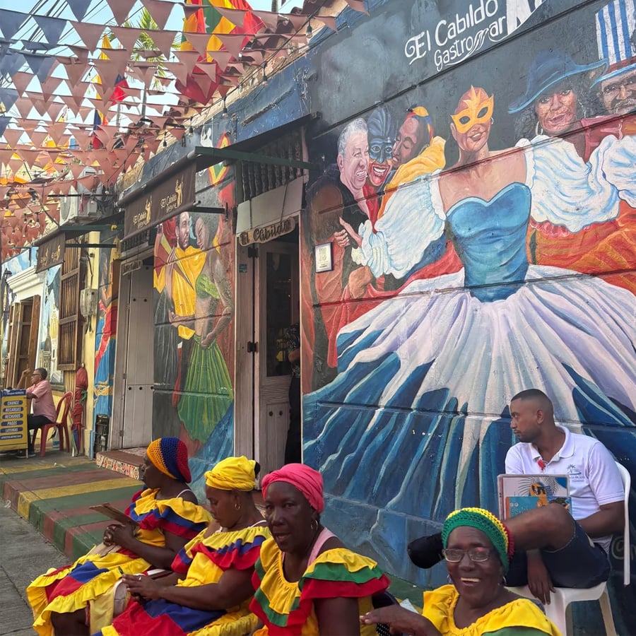 Four women in colorful dresses and headwraps sit on a sidewalk beside a man in a white shirt, in front of a vibrant mural and hanging flags on a lively street.