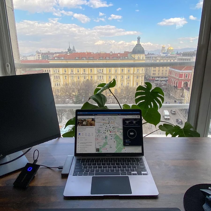 A laptop on a desk by a window shows a speed test and map. Next to it is a monitor and a plant. Outside, a cityscape with historic buildings and a blue sky with clouds is visible.