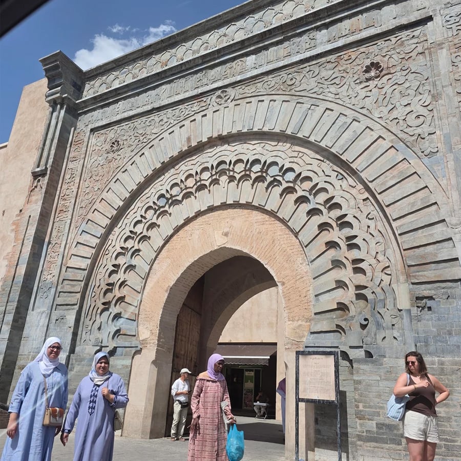 Three women stand near a large ornate arched gateway with intricate stone carvings and patterns. Other people walk nearby under a sunny blue sky. The architecture suggests a historic site.