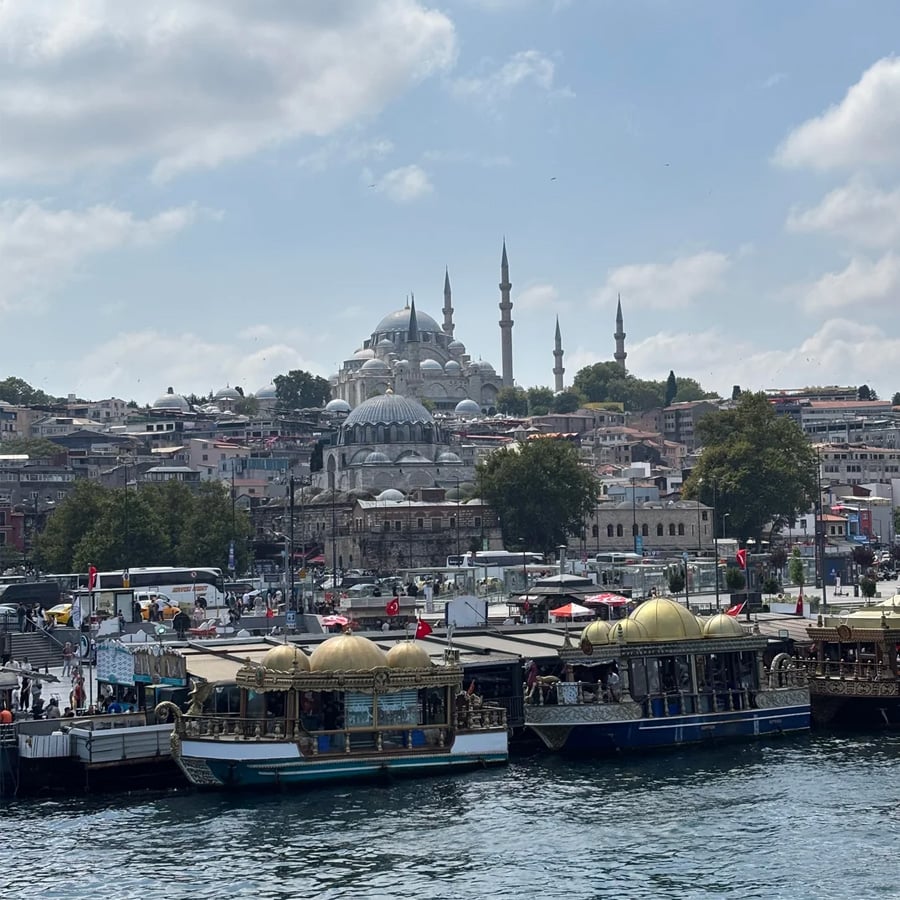 Boats docked by the waterfront with a busy market, trees, and historic mosques with multiple domes and minarets in the background under a partly cloudy sky.