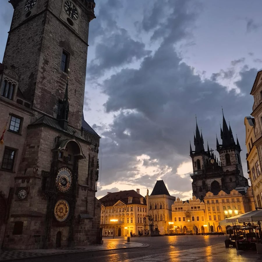 Twilight view of a historic town square with an ornate clock tower on the left, illuminated buildings, and a gothic church with spires in the background under a dramatic cloudy sky.