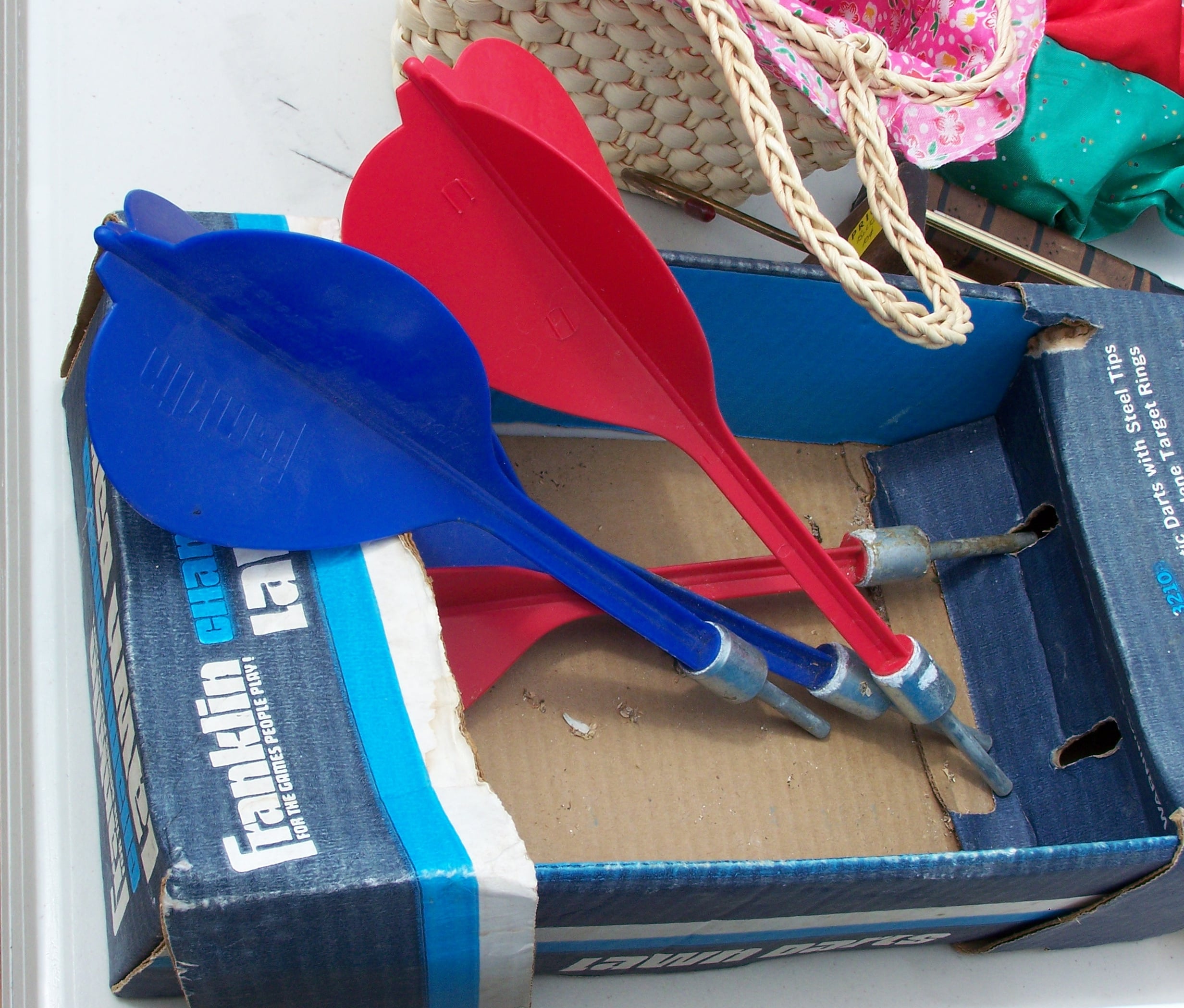 Three large lawn darts, two red and one blue, rest in a partially open cardboard box. A woven bag and some fabric are visible in the background.