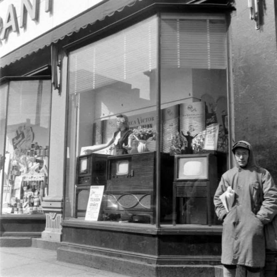 A person stands outside a storefront displaying several vintage televisions in the window. The shop has large glass windows, and the person is wearing a coat and cap, holding newspapers.