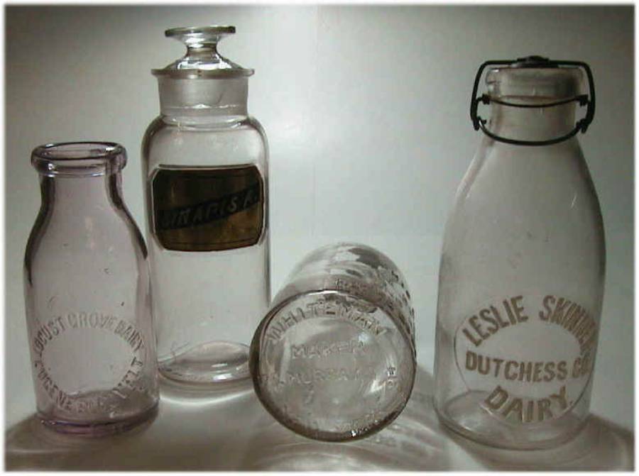 Four vintage clear glass bottles, including two milk bottles with embossed lettering, one lidded apothecary jar, and one bottle lying on its side, all displayed against a white background.