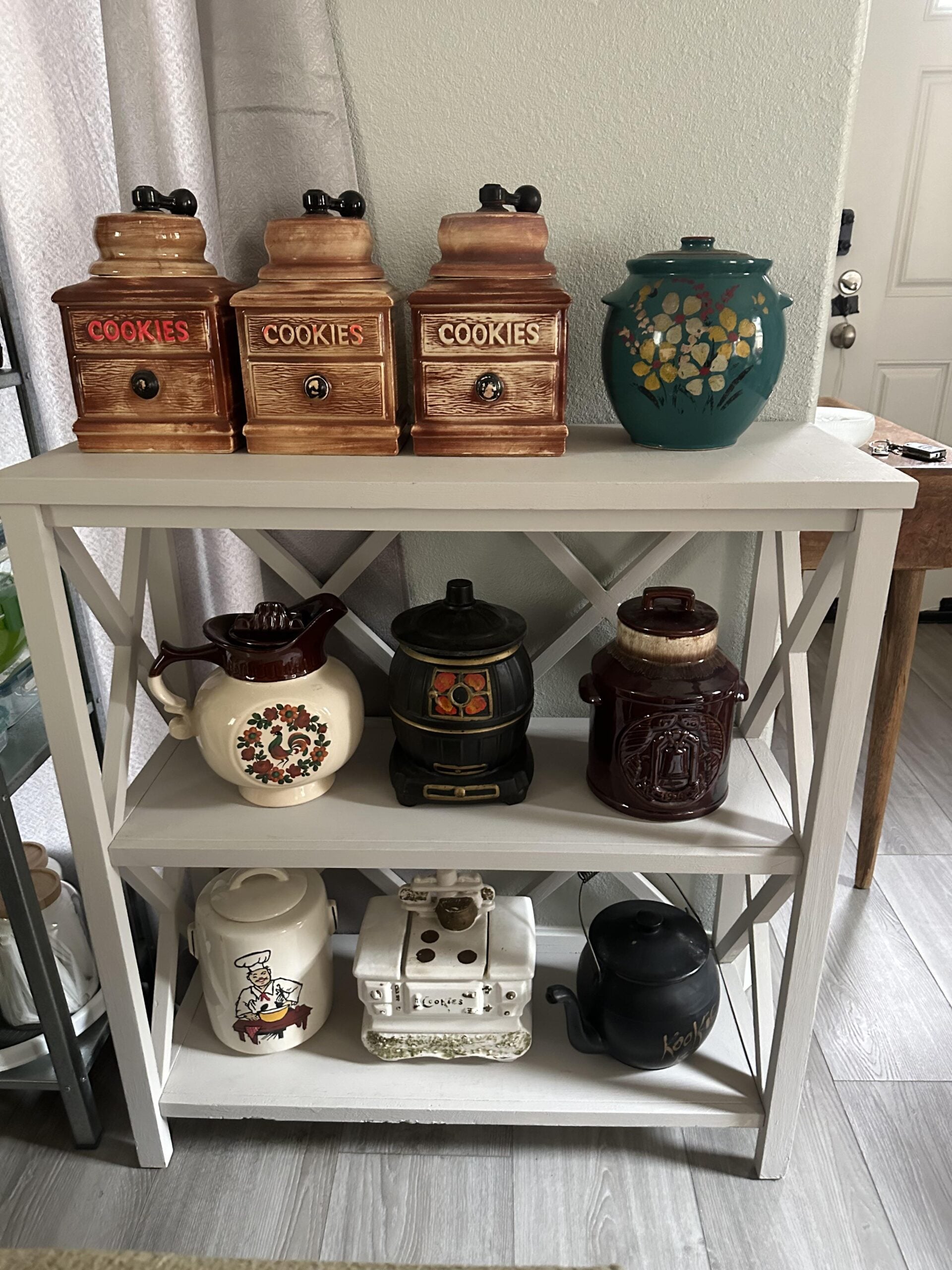 A white shelf holds three vintage cookie jars, a teal floral canister, and various decorative teapots and jars, all arranged neatly against a light wall near a door.