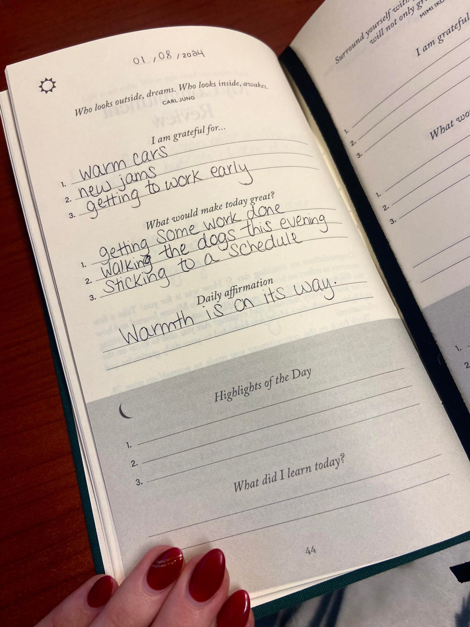 A hand with red nails holds open a journal filled out for January 8. Written entries include gratitudes, goals, a daily affirmation—“Warmth is on its way”—and blank spaces for highlights and lessons of the day.