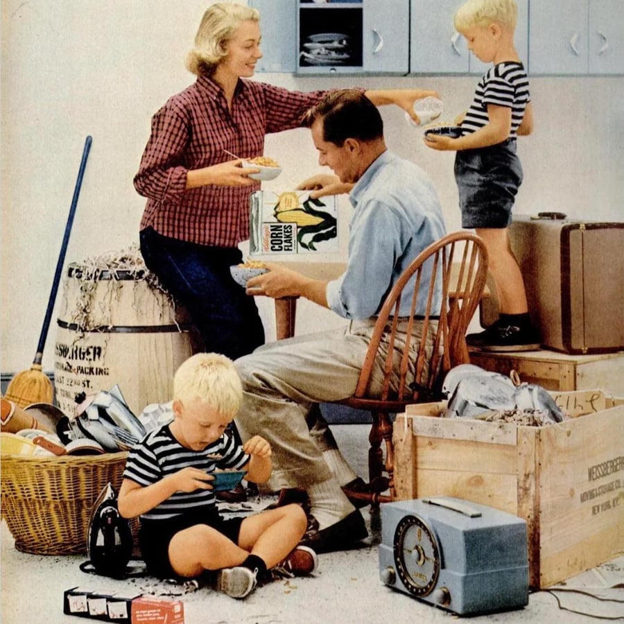A family of four eats in a cluttered room with boxes, bags, and appliances. The mother stands smiling, the father sits holding cereal, and two young boys in matching outfits eat on the floor and a crate.