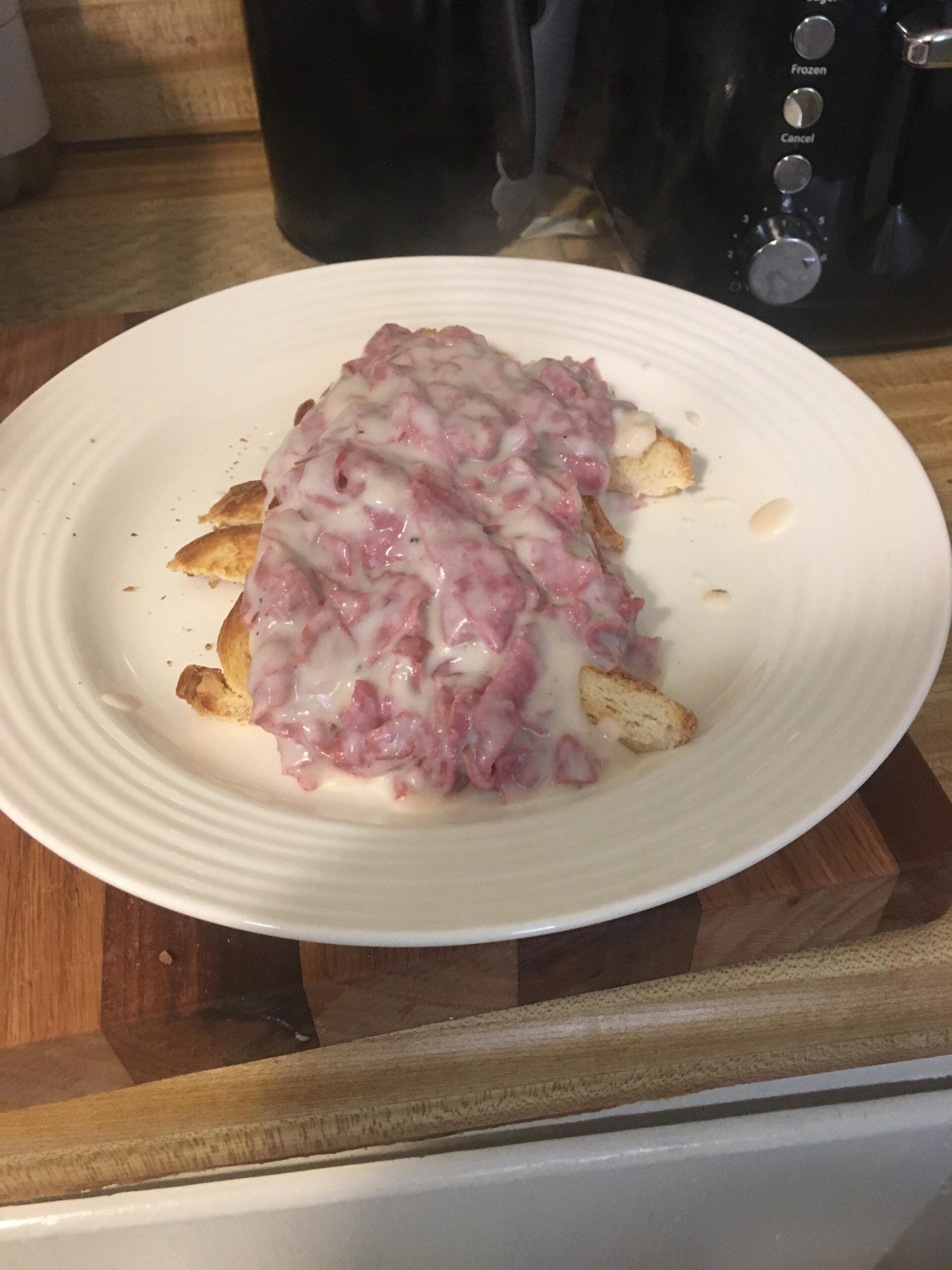A plate of toast topped with creamy chipped beef gravy sits on a wooden cutting board in a kitchen setting.