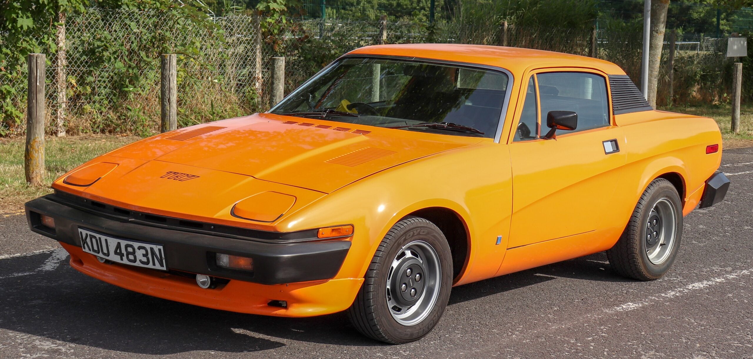 An orange classic sports car parked on a road, featuring pop-up headlights, a black roof section, and a UK license plate reading KDU 483N. Trees and a fence are visible in the background.