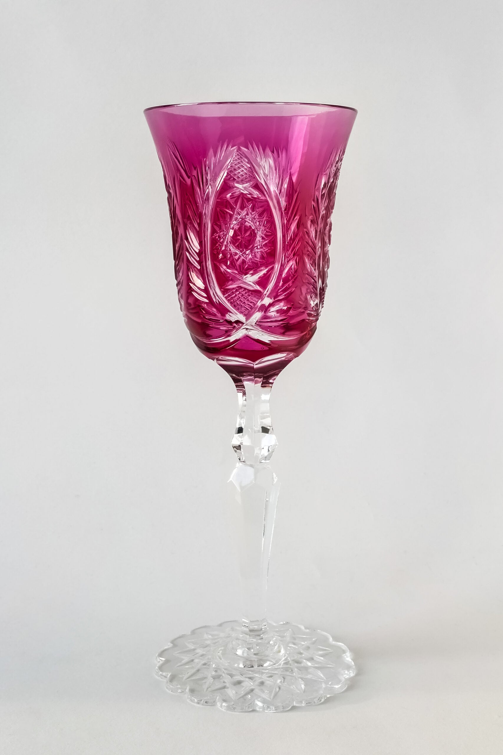 A crystal wine glass with an intricate cut design and a pink-tinted bowl, standing on a clear, faceted stem and base, against a plain light background.