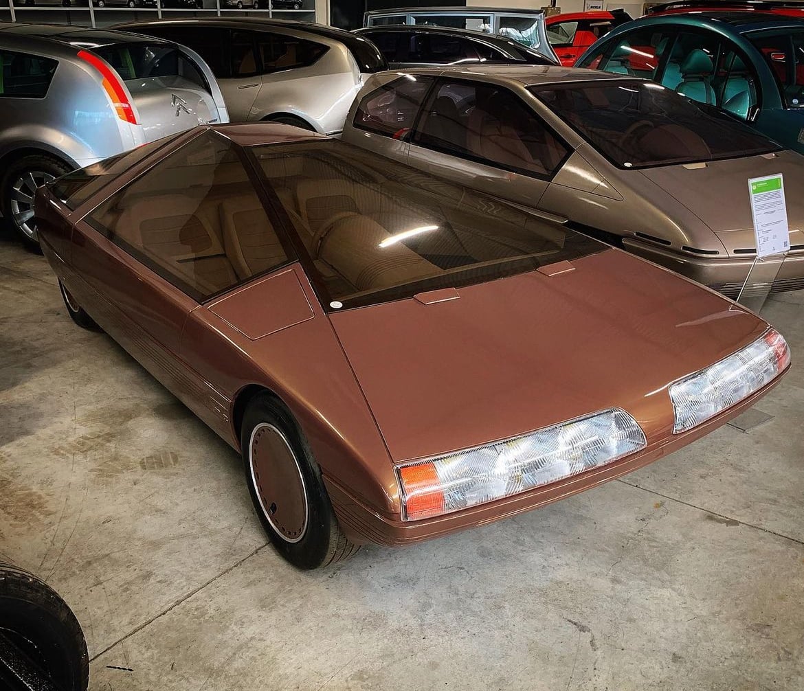 A vintage brown concept car with a sleek, angular design and large glass canopy is parked indoors among other unique vehicles.