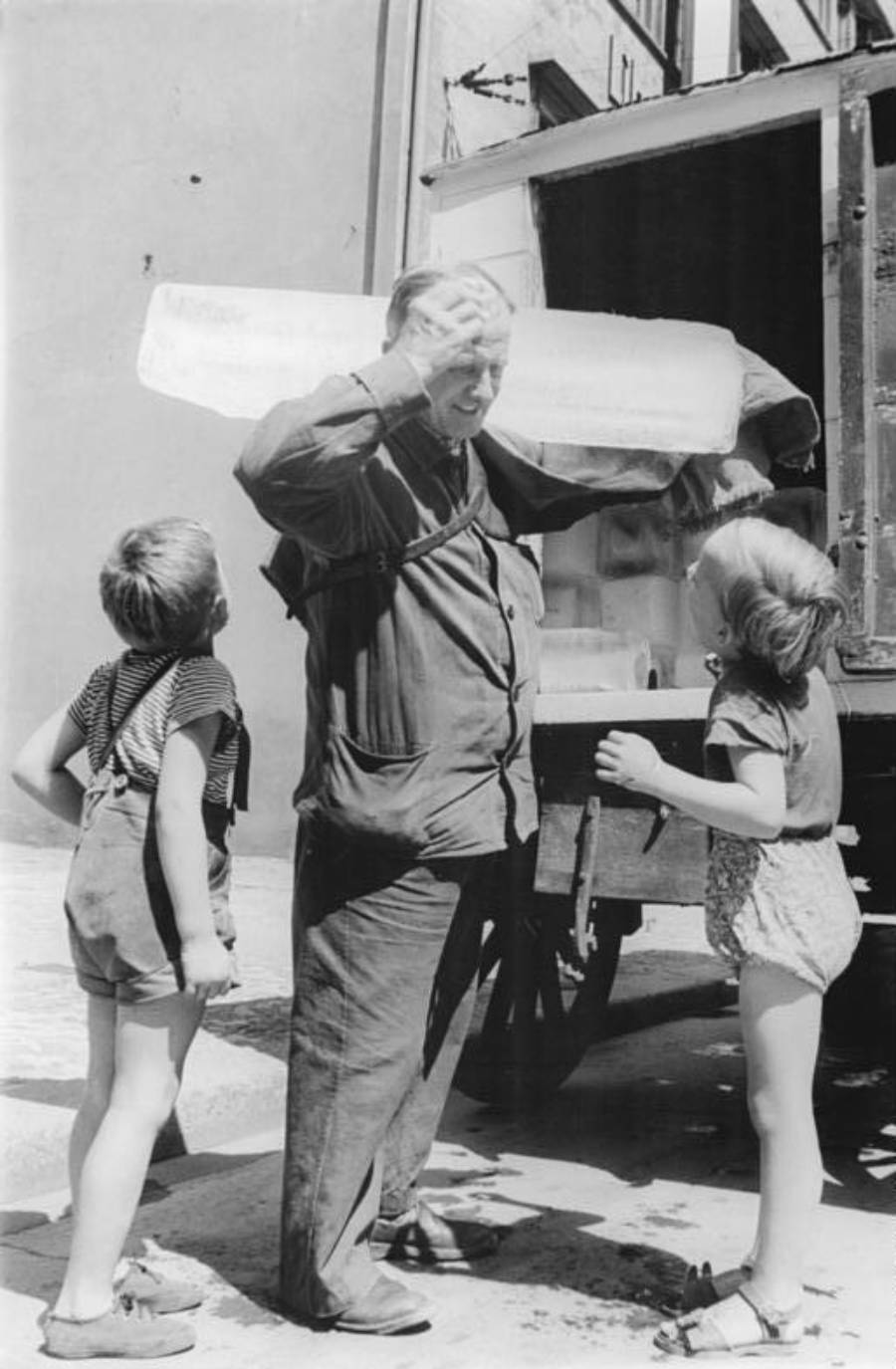 A man stands by a truck holding his head, with two children watching him and a large block of ice resting on his shoulder.