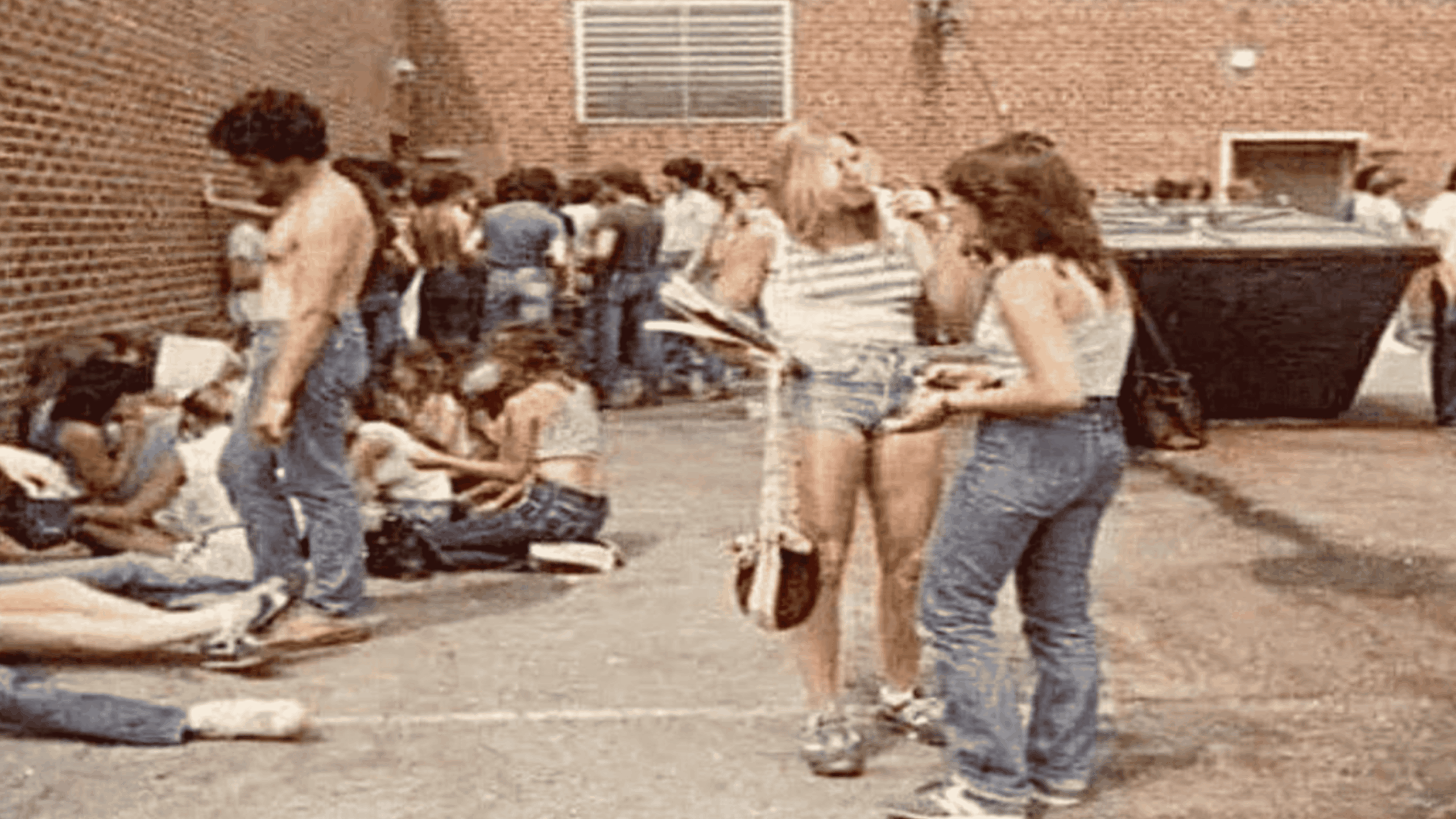A group of teenagers in casual 1970s clothing hangs out in a brick-walled alley, some sitting, some standing and talking, near a large dumpster.